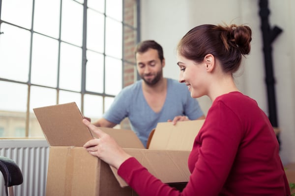 Attractive young couple moving house getting ready to unpack a cardboard carton of personal possessions below a big window