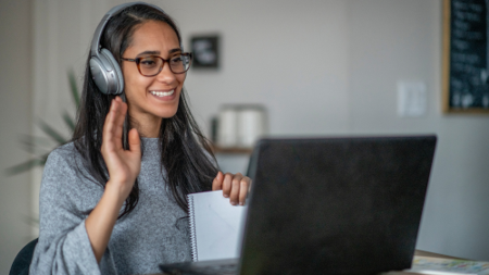 Woman wearing headphones smiling and waving at laptop during language training session