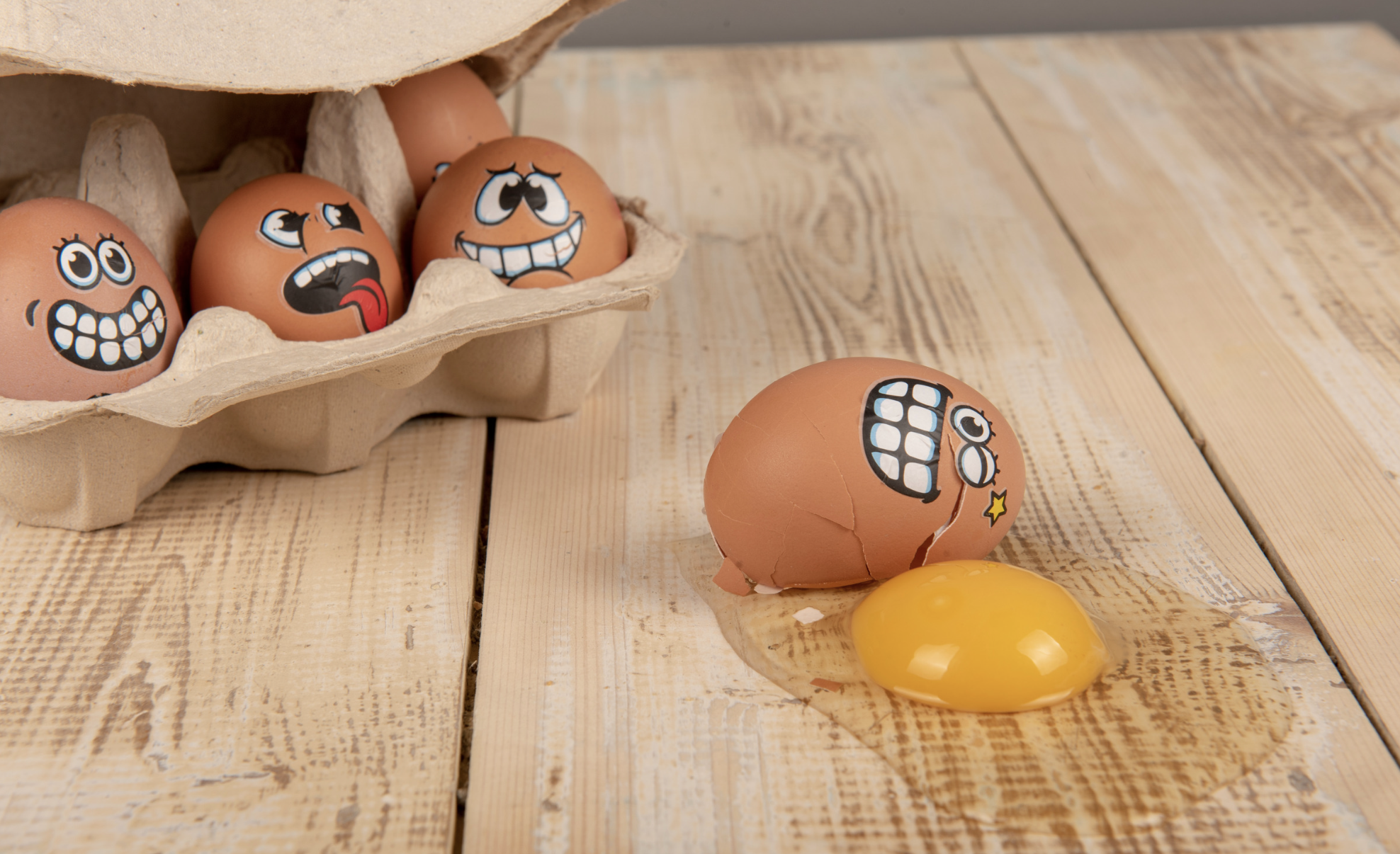 A cartong of eggs with comical faces with different expressions painted on them. One of the eggs is laying outside the carton, cracked with yolk running out, and with a nervous expression