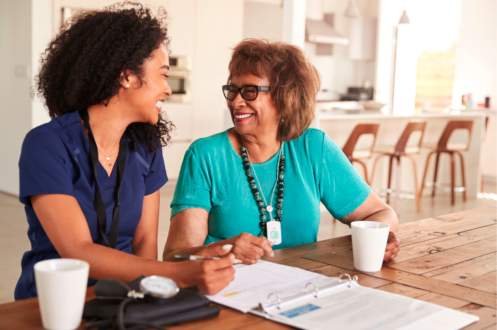 A nurse sitting at a table with an older female patient gathering patient data. They are smiling and having a pleasant exchange.