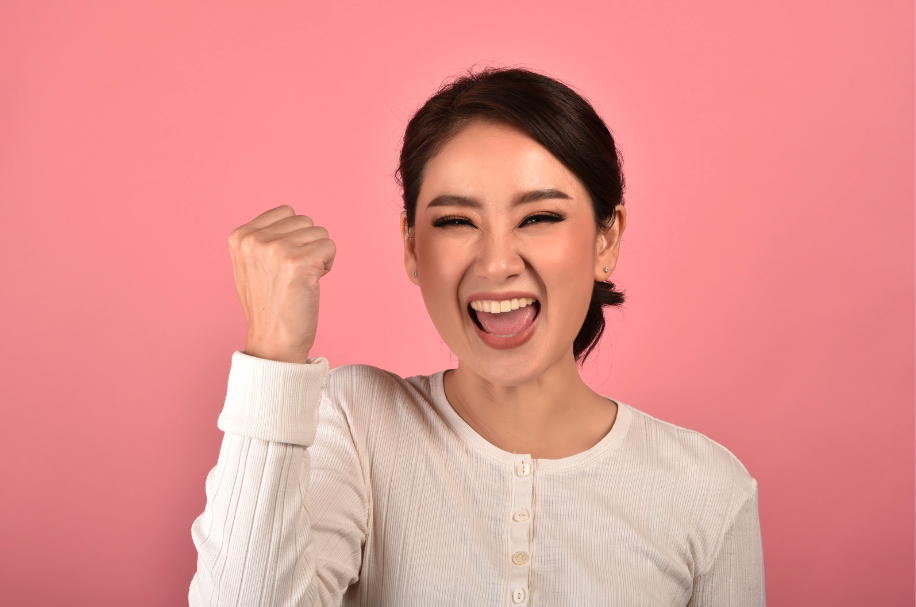 Young woman against a pink background, holding up a fist feeling happy and triumphant 