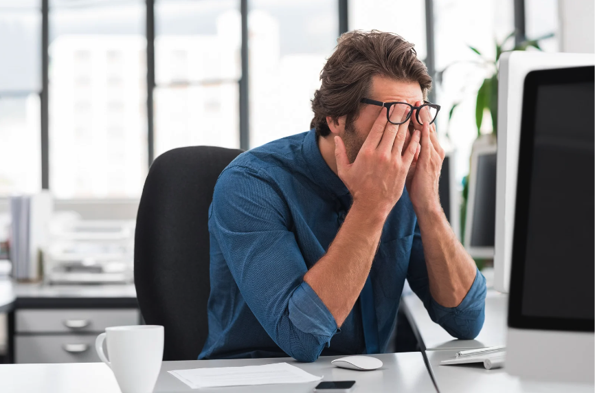 Man sitting at desk, rubbing his eyes underneath his eyeglasses in frustration.