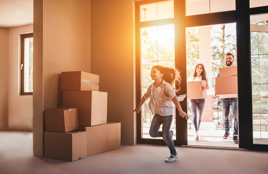 Family who has relocated. The young girl is running into her new home. Mother and father are following closely behind her, carrying moving boxes.