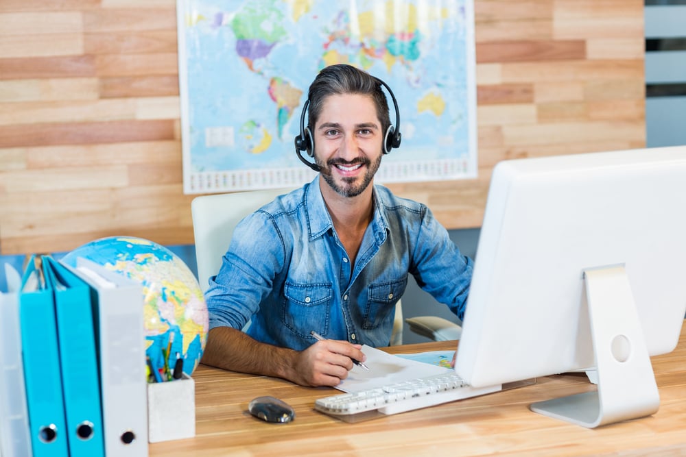 Male travel agent sitting at his desk wearing a headset using his new bilingual skills from language training