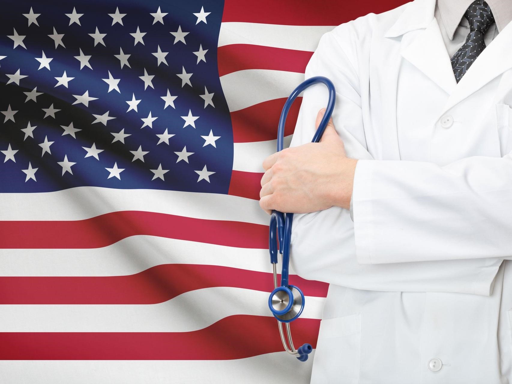 Doctor in white coat holding stethoscope standing in front of an American flag to showcase language training for relocation